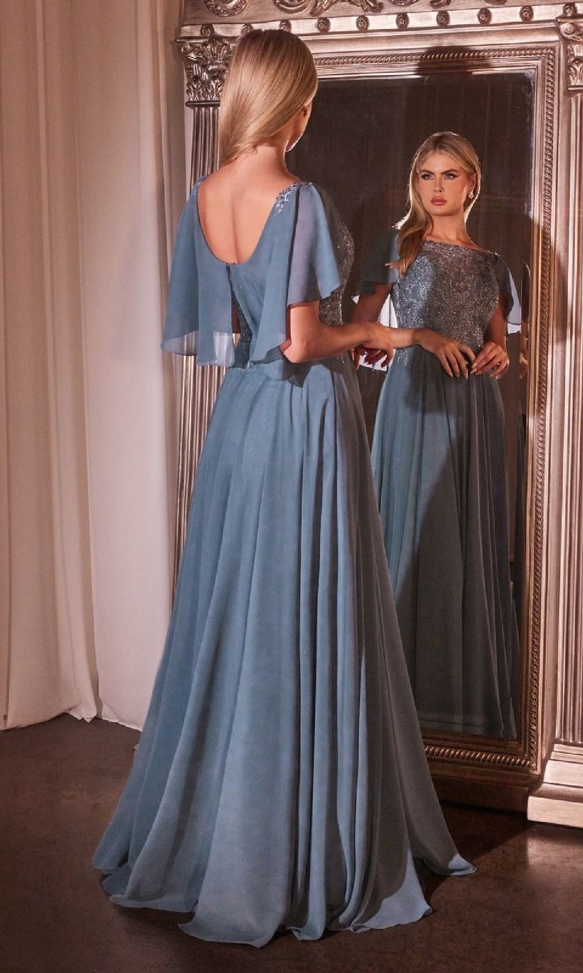A woman in the Cape-Sleeve Long A-Line Prom Dress: Ladivine HT101 stands before a large ornate mirror, her flowing blue gown and elegant cape sleeves beautifully reflected.